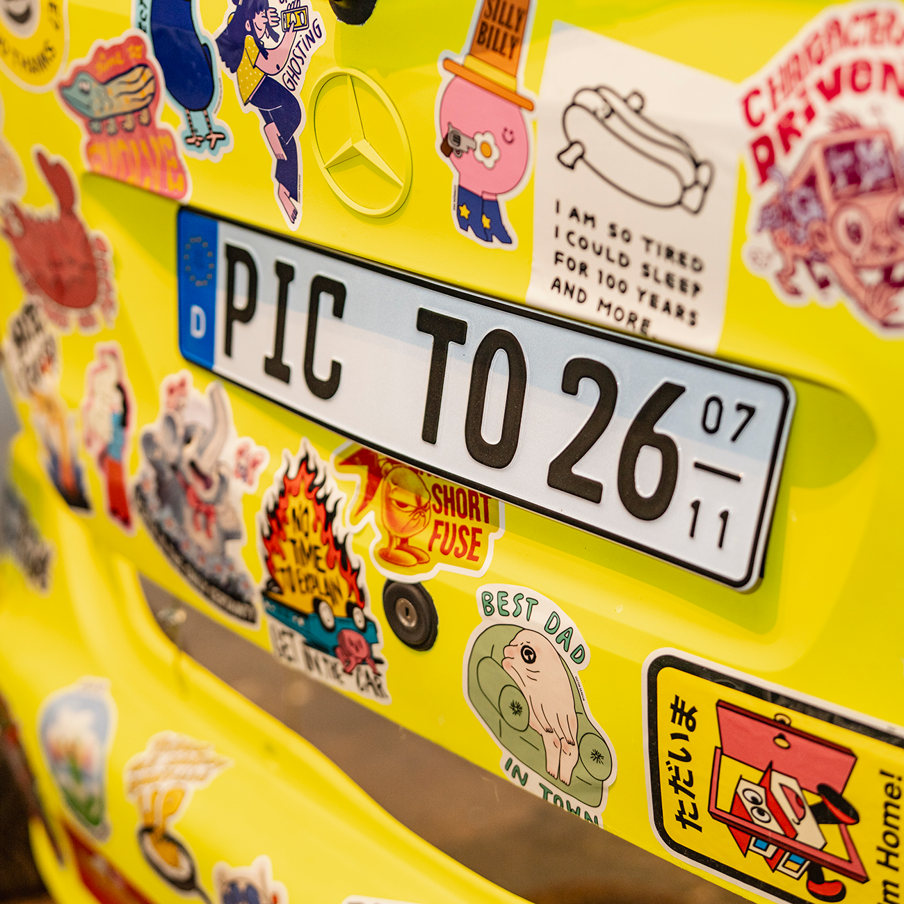 Yellow Mercedes body covered in bumper stickers in various shapes and sizes, with a german licence plate.