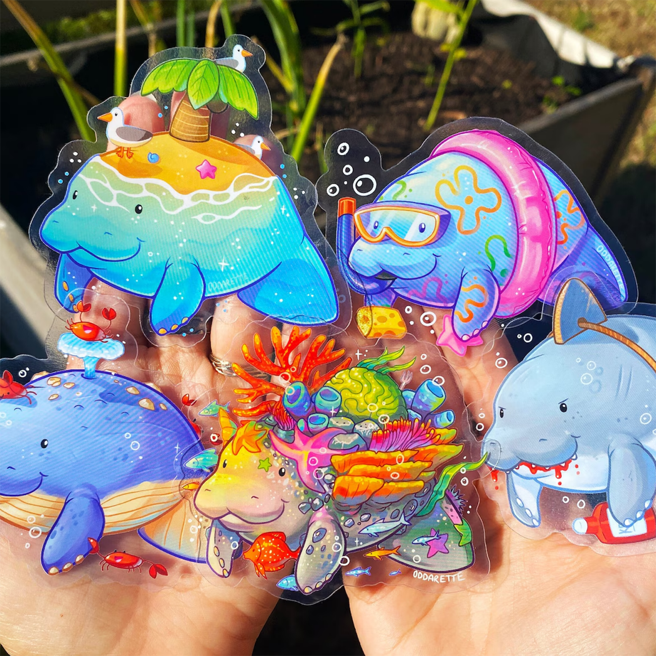 Hand holding five manatee stickers, with different summery ocean vibes, held against green plants and a planter.