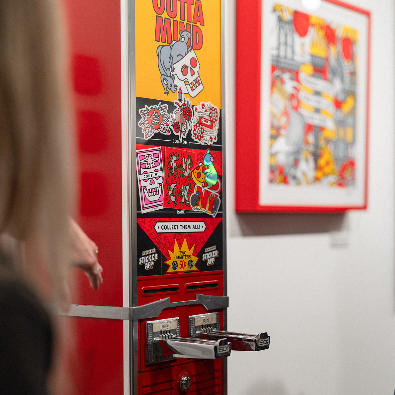 A person uses a vintage sticker vending machine with colorful skull-themed designs in an art gallery setting.