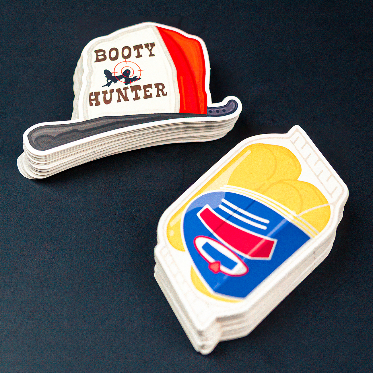 Two vinyl die-cut stickers showing two loafs of bread in a plastic container, and a cap with the text "booty hunter" on a dark background.