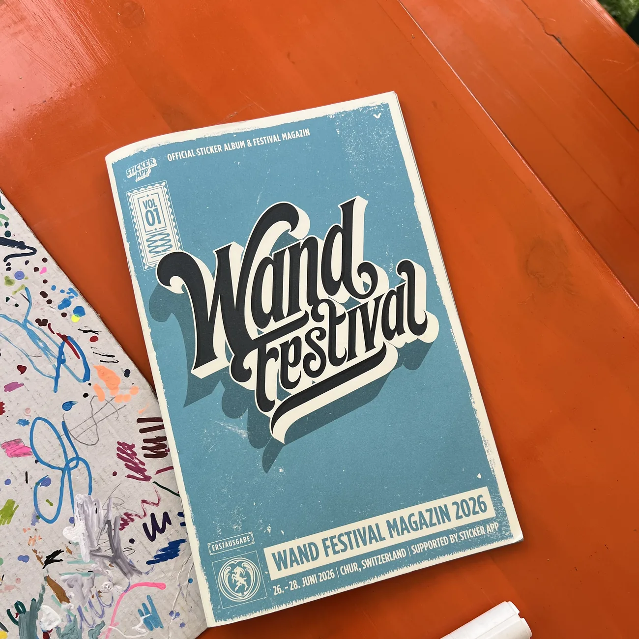 StickerAppが協賛するWand Festival 2026の公式ステッカーアルバム兼フェスティバルマガジンと、オレンジのテーブルに置かれたペイントマーカー