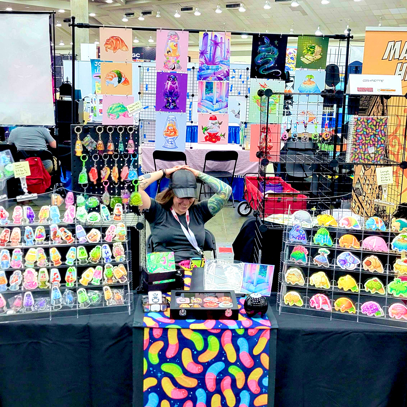 A booth displaying colorful art prints, stickers, and keychains at a convention. Oddarette sits behind the table adjusting their cap.