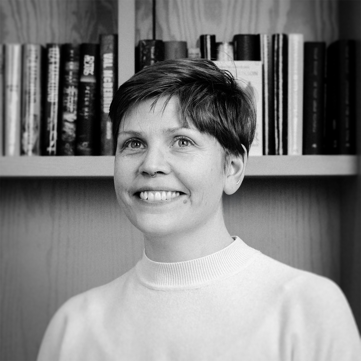 Person smiling in front of a bookshelf with neatly arranged books. Black and white image with a wooden background.