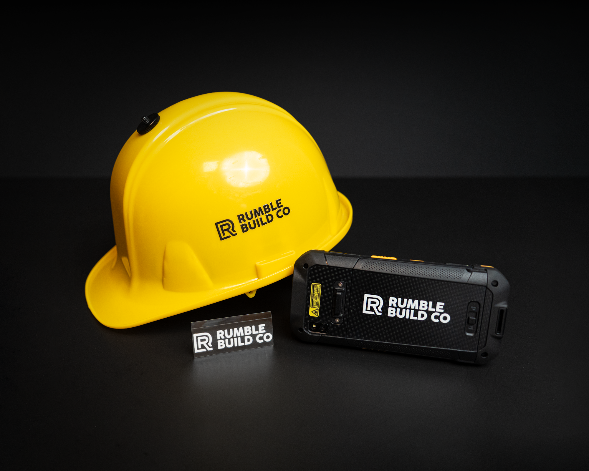 Yellow hard hat, rugged smartphone, and transfer sticker with "Rumble Build Co" logo on a black background.