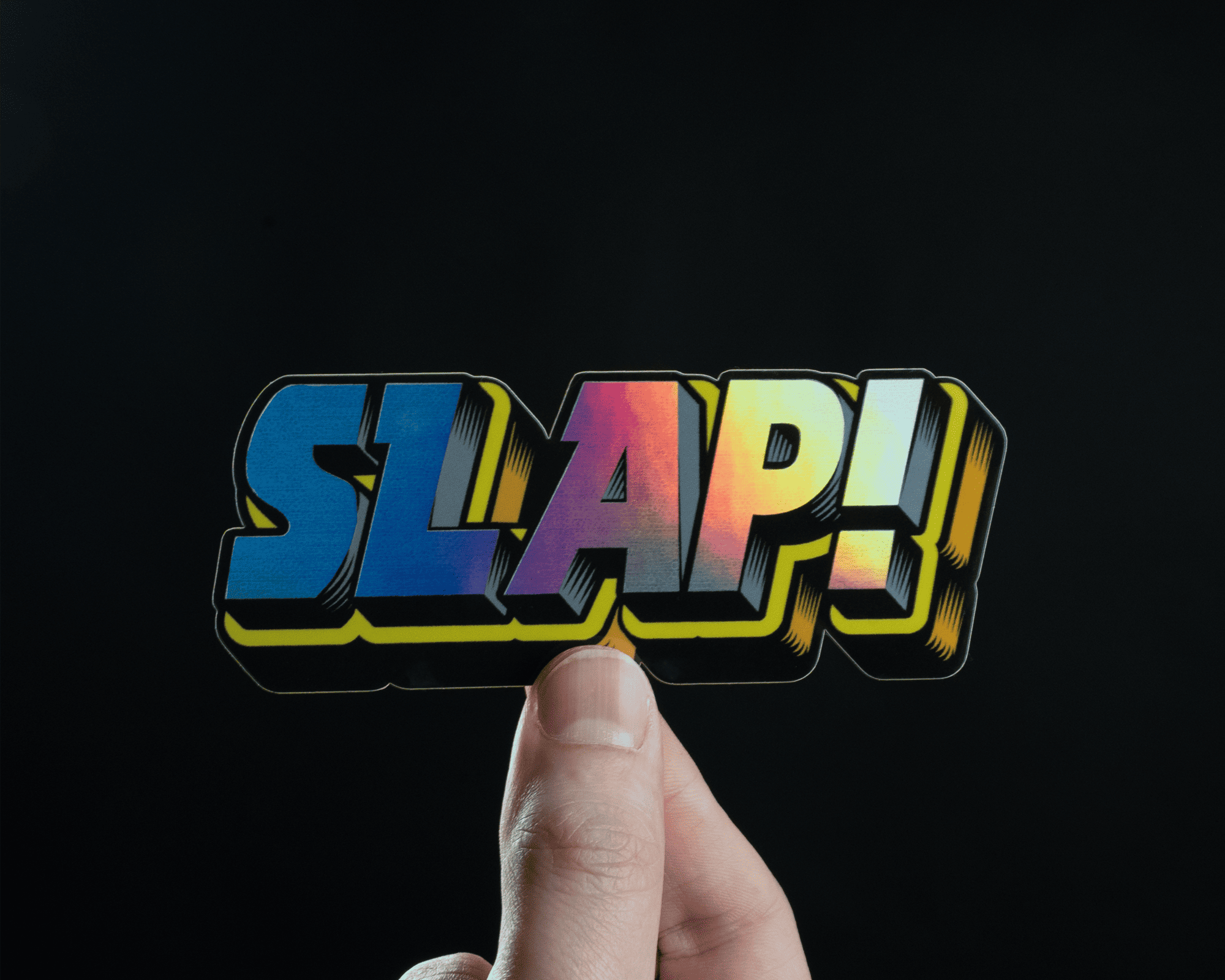 Hand holding a mirror sticker with the word "SLAP!" in bold, stylized letters against a dark background.