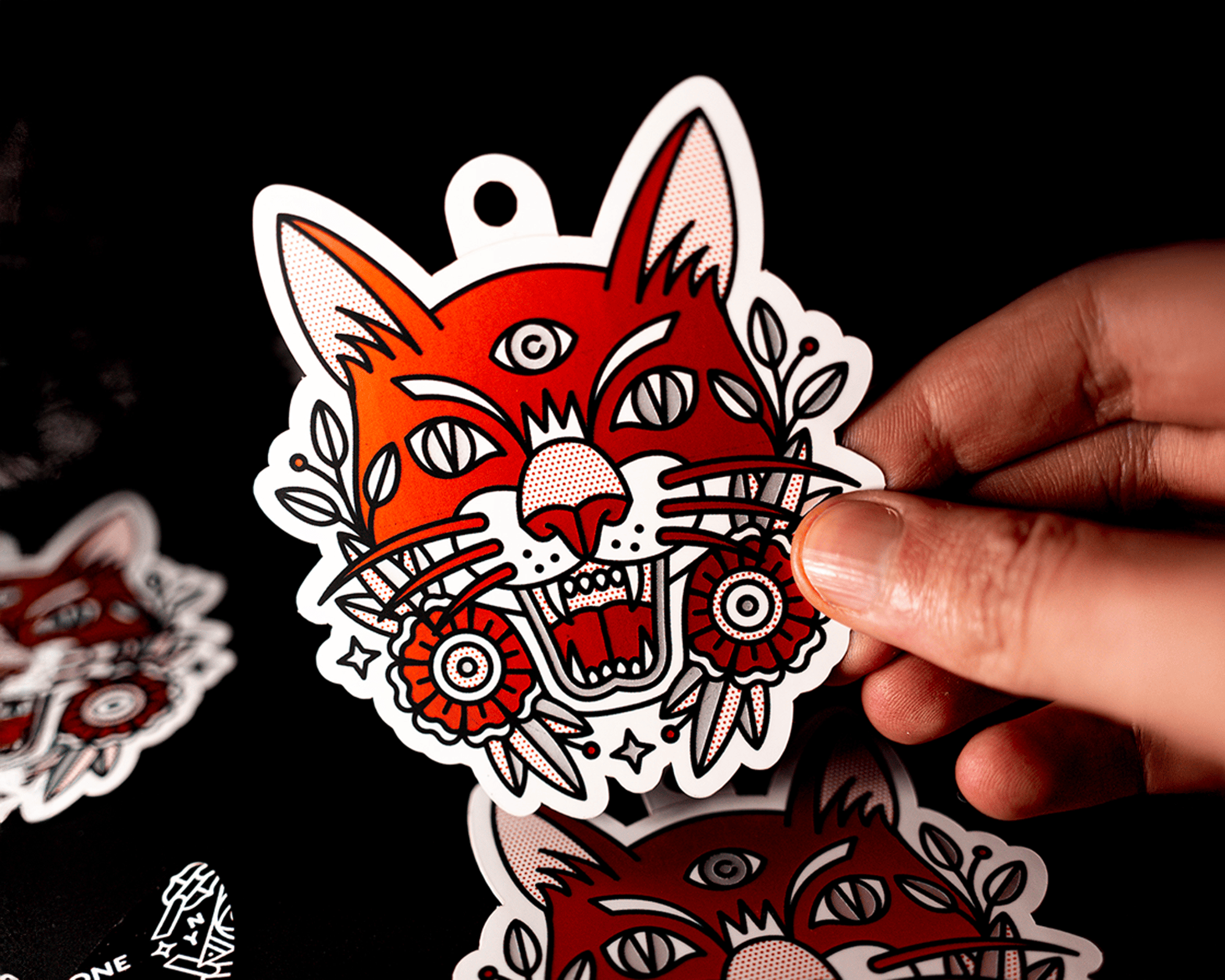 Hand holding a hang tag sticker of a stylized red cat with three eyes, open mouth, and floral decorations on a black background.
