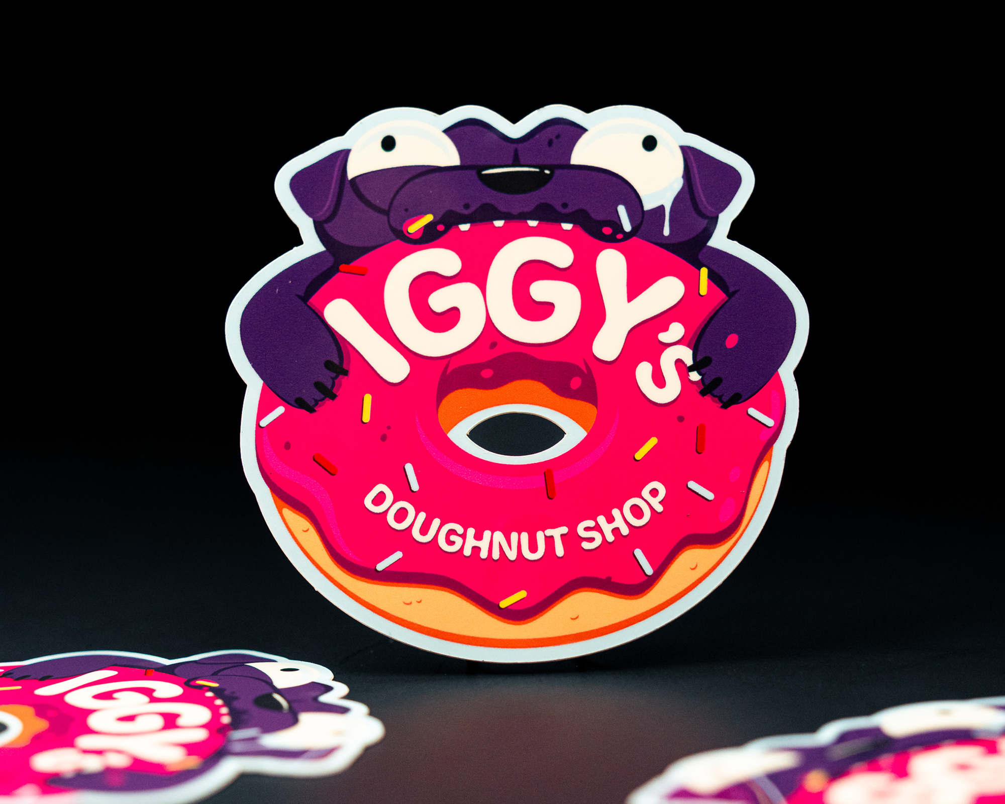 Sticker of cartoon pug with a pink donut labeled "Iggy's Doughnut Shop," surrounded by sprinkles, on a dark background.