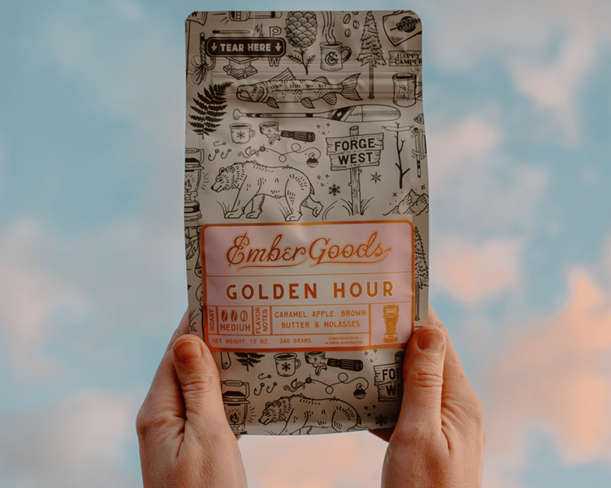空を背景に、メタリックラベルを貼ったEmber Goods Golden Hourのコーヒー袋を持つ手