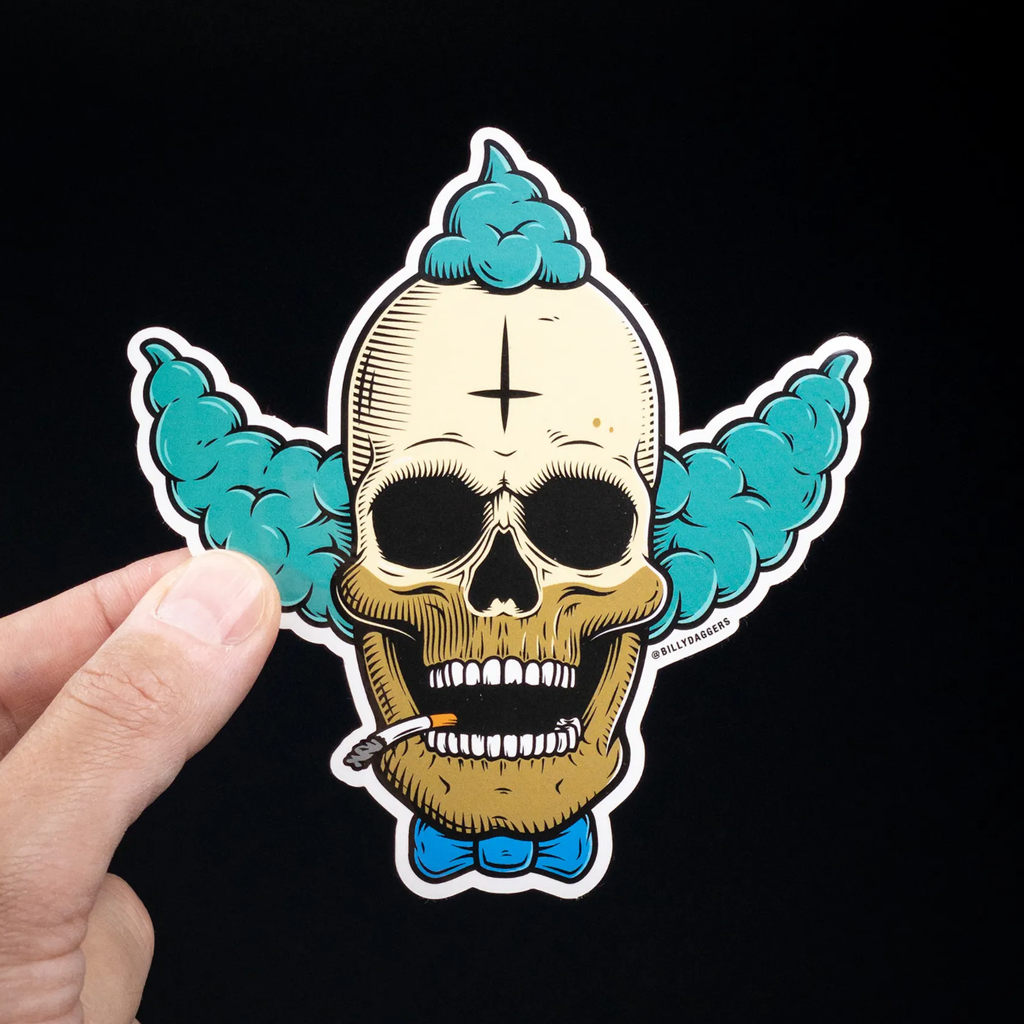 Hand holding a sticker of a skull with clown features, blue hair, and a cigarette, set against a black background.