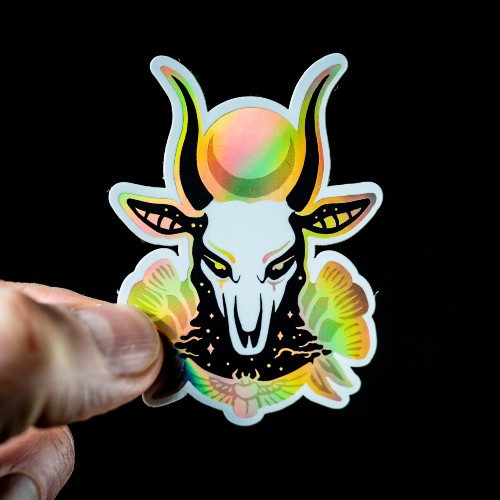 A hand holding a custom holographic sticker with a evil devil skull on it