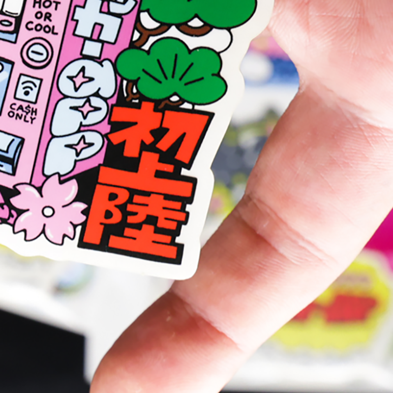 Close-up of a hand holding a colorful sticker featuring Japanese characters, decorative elements, and the word "HAPPY" in bold letters.